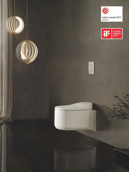 A GROHE Sensia Arena shower toilet attached to a dark grey wall above a shiny, black floor, with two lamps hanging from the ceiling.