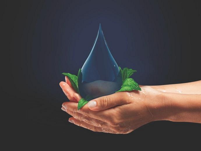 Hands gently cupping a large, glowing blue water droplet surrounded by green leaves against a dark background. The image conveys concepts of environmental conservation, purity, and the importance of water resources.